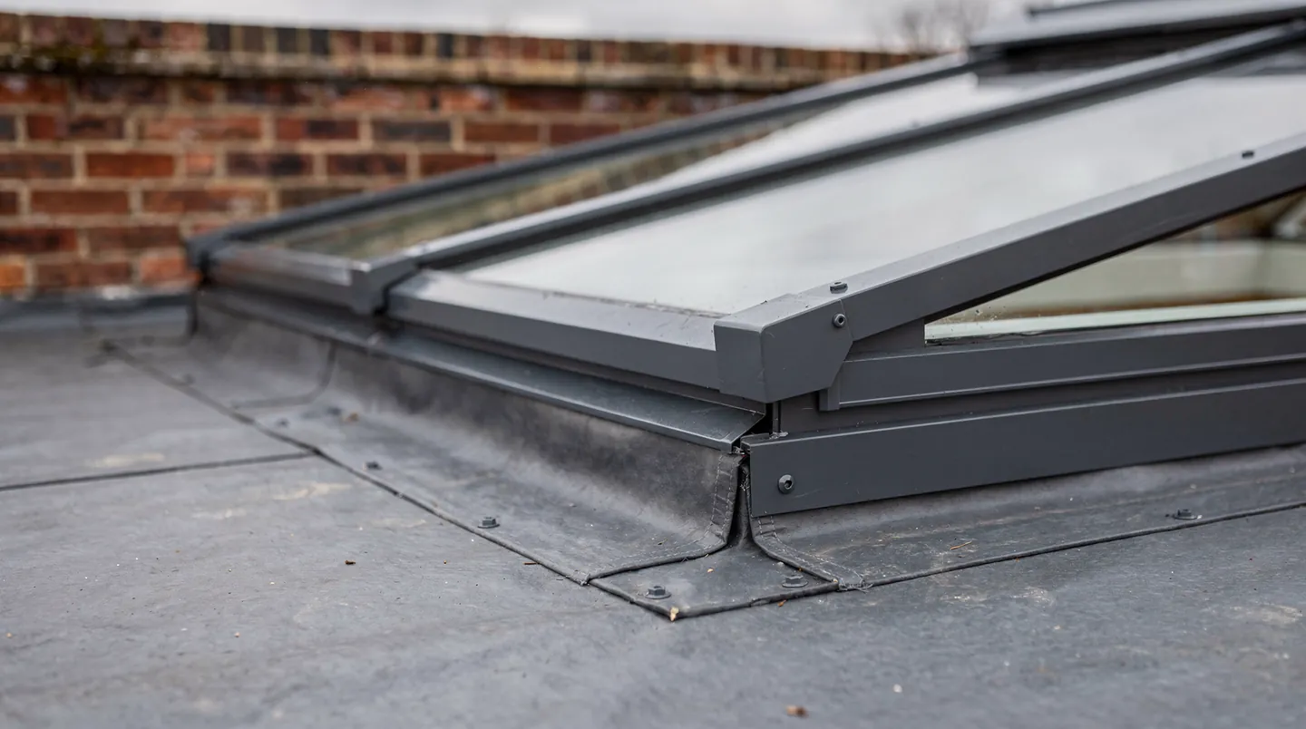 The corner of a dark grey roof lantern on its kerb on a flat roof, with the membrane dressed up the side of the upstand